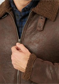 Uomo che sta chiudendo una giacca di pelle marrone con colletto e polsini in shearling, indossando una camicia a quadri blu sotto.