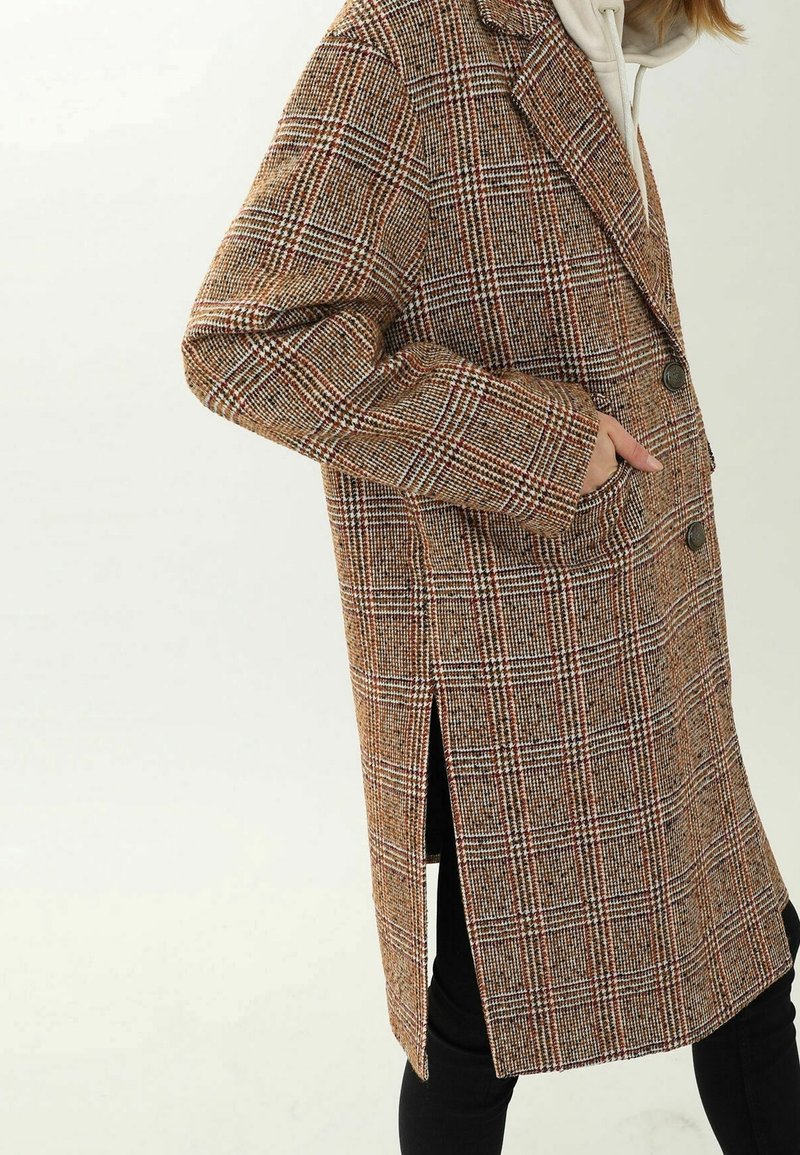 Tarkistettu villakangastakki ruskean sävyissä, jossa on yksirivinen malli, kaksi nappia, sivutaskut ja sivuhalkiot liikkuvuuden helpottamiseksi.