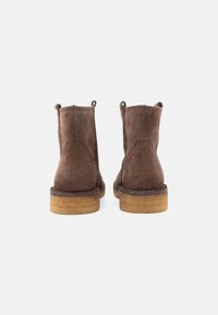 Botas de tornozelo em camurça castanha com solas de crepe, vistas de trás sobre um fundo claro e simples.