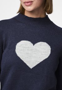 Pull bleu marine en maille avec un motif cœur blanc sur la poitrine. Il présente un col rond côtelé et une surface texturée.