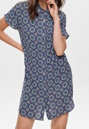 Kvinde iført kjole med korte ærmer og knapper foran med et blåt cirkulært blomsterprint, poserende med den ene hånd på hoften og den anden nær ansigtet.