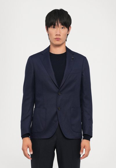 Blazer azul marino con un diseño estructurado, dos bolsillos frontales, botones negros y detalles de costura sutiles. Lleva encima un suéter oscuro.