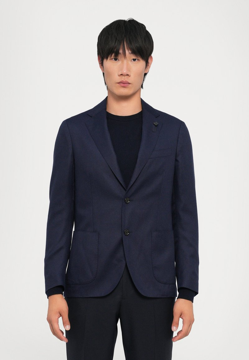 Navy blazer in a structured design with two front pockets, black buttons, and subtle stitching details. Worn over a dark sweater.