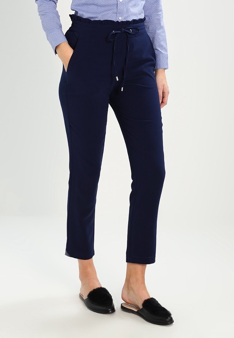 Femme portant un pantalon bleu marine fuselé avec taille à cordon et des chaussures à enfiler noires ornées de fourrure, debout avec une main dans la poche.