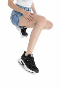 Zapatillas de malla negra con suelas blancas. Incluyen características como cordones, acentos de logo y un diseño acolchado. El modelo lleva puesta una falda de mezclilla.