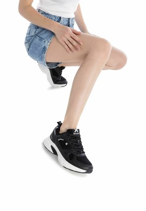 Zapatillas de malla negra con suelas blancas. Incluyen características como cordones, acentos de logo y un diseño acolchado. El modelo lleva puesta una falda de mezclilla.