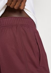Burgundy színű sportos rövidnadrág, elasztikus derékrésszel, textúrált anyagból, oldalsó zsebbel és laza szabással, félig látható világos háttér előtt.