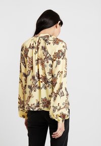 Yellow blouse with floral paisley pattern, long sleeves, and buttoned cuffs. Soft fabric drapes gently over the shoulders and back.