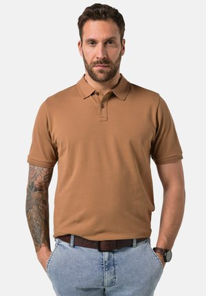 Mężczyzna z brodą, z tatuażem na lewej ręce, ubrany w brązową koszulkę polo, niebieskie dżinsy, brązowy pasek i zegarek, stoi z rękami w kieszeniach na białym tle.