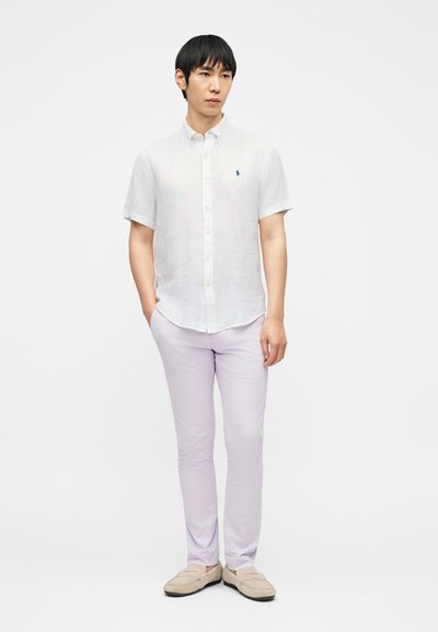 Νεαρός άνδρας με μαύρα μαλλιά, φορώντας λευκό πουκάμισο με κοντά μανίκια και κουμπιά και ανοιχτόχρωμο λιλά παντελόνι, στέκεται με τα χέρια στις τσέπες.