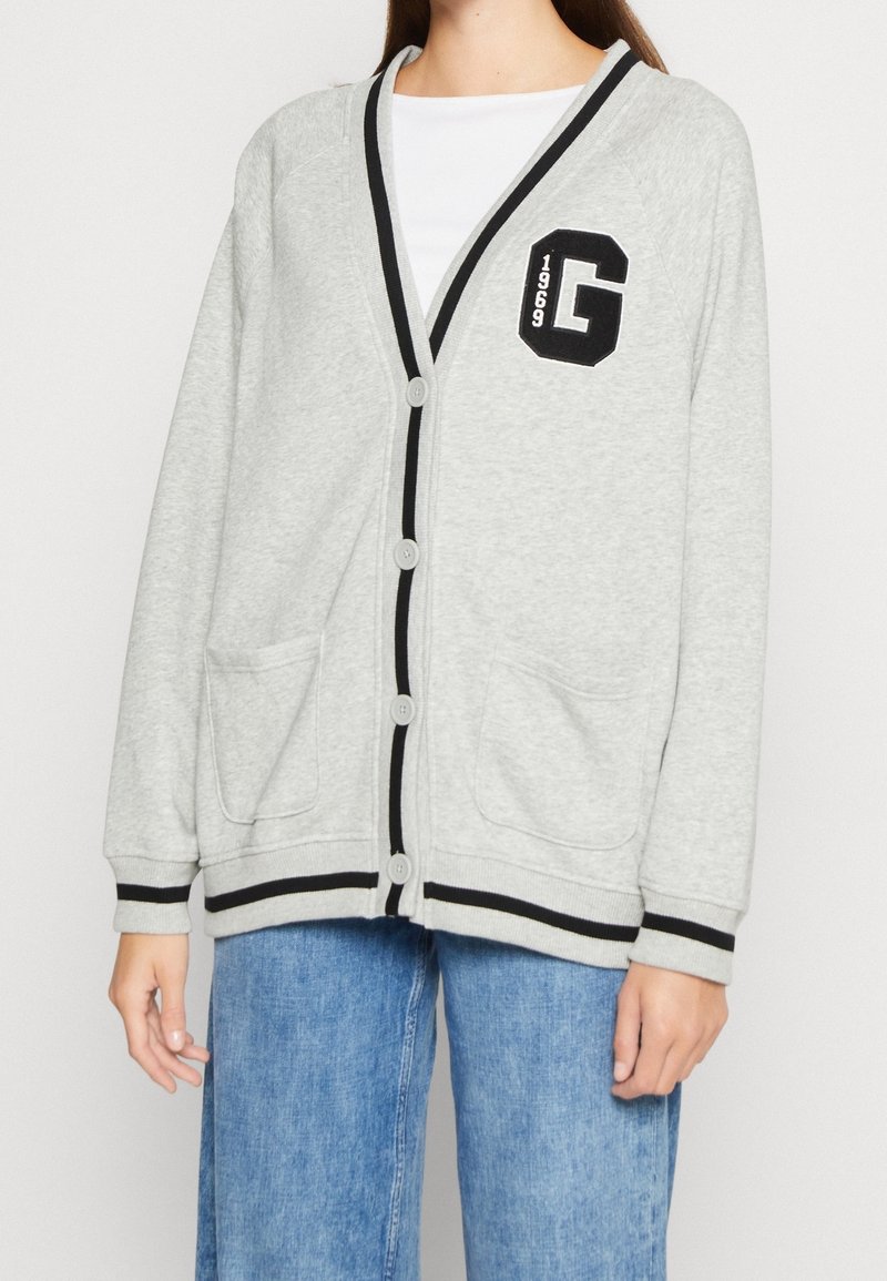 Personne portant un cardigan gris clair avec bordure noire, boutons, écusson avec "G" et "1969," et un jean bleu.