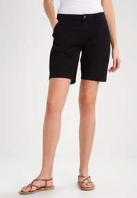 Kvinna i svarta knälånga shorts, vit topp och bruna flator, står med ena handen i fickan mot en ljus bakgrund.