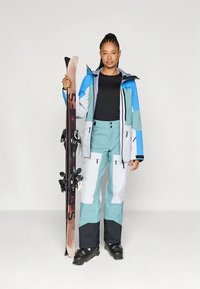 Skikleding bestaande uit een blauwe, witte en grijze jas; teal broek; houdt zwarte en roze ski's vast; waterdicht materiaal; met ritssluitingen voor de zakken.