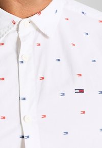 Camisa blanca de botones con un patrón de pequeños cuadros rojos y azules. Incluye un pequeño logotipo bordado en el pecho. Textura suave.