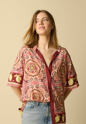 Jeune femme aux cheveux blonds mi-longs porte une chemise ample à motifs boutonnée et un jean bleu clair, debout devant un fond beige.