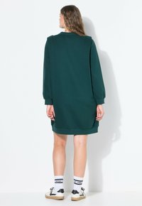 Grünes Sweatshirtkleid mit langen Ärmeln, gerippten Bündchen und seitlichen Schlitzen. Weicher, strukturierter Stoff, knielang, kombiniert mit weißen Socken und Turnschuhen.