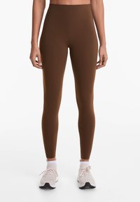Barna, magas derekú leggings, amelyek testhez simuló kialakításúak és sima textúrájúak, világos színű sportcipőkkel és fehér bokazoknival párosítva.