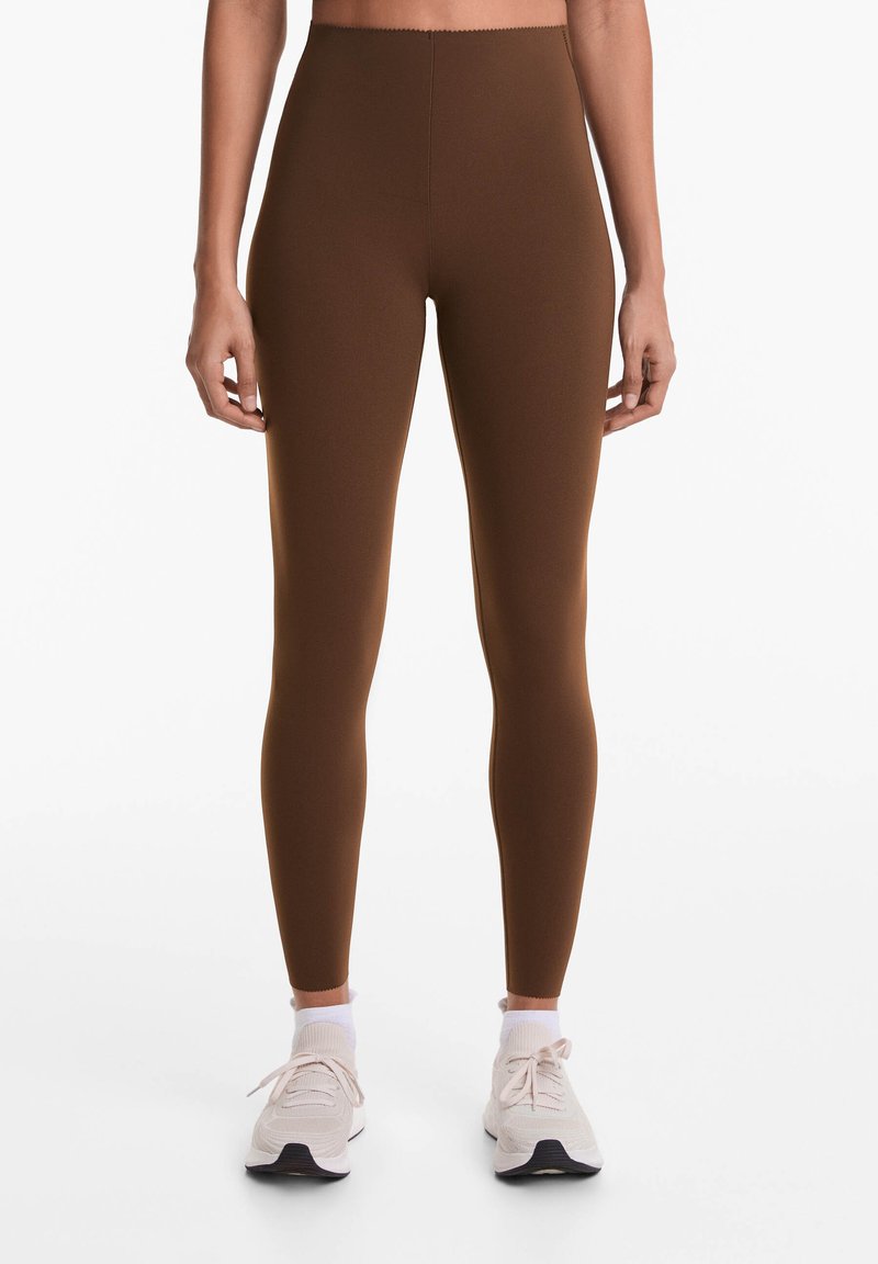 Barna, magas derekú leggings, amelyek testhez simuló kialakításúak és sima textúrájúak, világos színű sportcipőkkel és fehér bokazoknival párosítva.