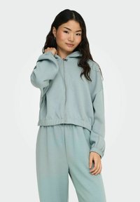 Mintgrønn cropped hettegenser med glidelås, elastiske mansjetter og midjebånd. Stoffet virker mykt og glatt, og passer til matchende bukser.