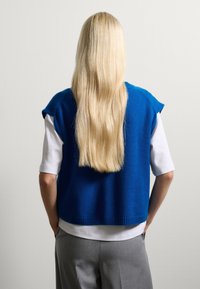 Blauwe gebreide vest met mouwtjes, gelaagd over een wit shirt met korte mouwen; gecombineerd met grijze broek; zachte textuur en ontspannen pasvorm.