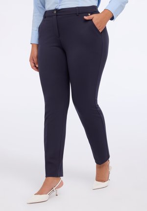 Kvinde iført marineblå slim-fit ankellange bukser med frontlommer og lyseblå skjorte med lange ærmer, kombineret med hvide slingback-hæle.