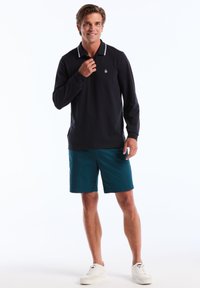 Chemise polo à manches longues marine avec des accents de col blanc, assortie à un short teal et des baskets blanches, avec un logo brodé sur la poitrine.