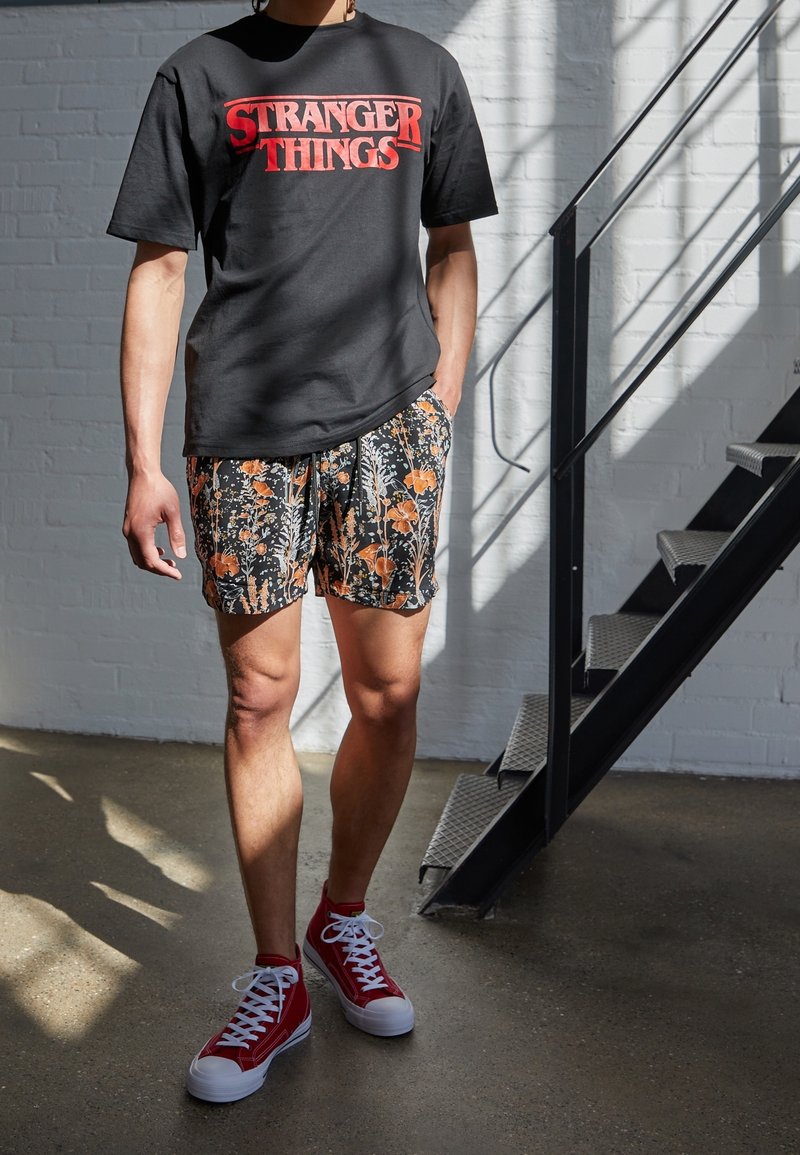 Personne portant un t-shirt noir de Stranger Things, un short à fleurs et des baskets montantes rouges, se tenant à l'intérieur près d'un escalier en métal.