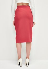 Jupe crayon rouge à taille haute, en tissu texturé, avec une couture design courbée. Portée avec des escarpins blancs à bout fermé, mettant en valeur une silhouette épurée.