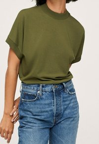 Oliwkowa bluzka z krótkim rękawem i prążkowanym okrągłym dekoltem, w połączeniu z jasnoniebieskimi spodniami o wysokim stanie z zapięciem na zamek i guzik.