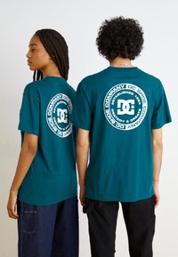 Teal bomulls t-skjorter med korte ermer, med et sirkulært hvitt grafisk logo på ryggen. Bærers av to personer som snur ryggen til.