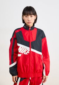 Rdeča in črna vetrovka z belimi akcenti; ima visok ovratnik, celoten zadrgo in velik Nike logotip spredaj. Tekstura tkanine.