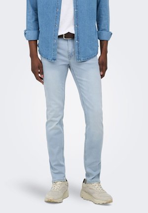 Homme portant un jean slim bleu clair, des baskets beige, un t-shirt blanc et une chemise en jean bleue avec les manches retroussées, debout devant un fond blanc.