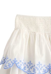 Hvid bomuldsnederdel med rynket, elastisk talje. Nederste kant har blå blomsterbroderi og en scallop-kant.