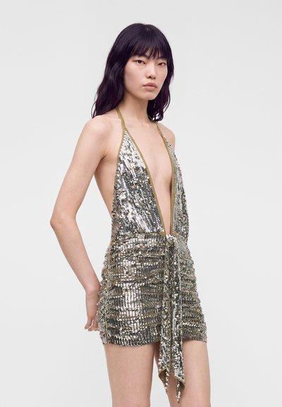 Femme aux longs cheveux noirs portant une mini-robe halter argentée à sequins avec un profond décolleté en V et un détail noué à l'avant.