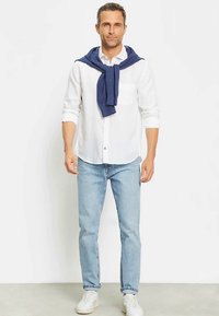 Camicia bianca a maniche lunghe, jeans azzurri chiari e scarpe da ginnastica bianche. Maglione blu scuro appoggiato sulle spalle. Stile casual, tessuti leggeri.