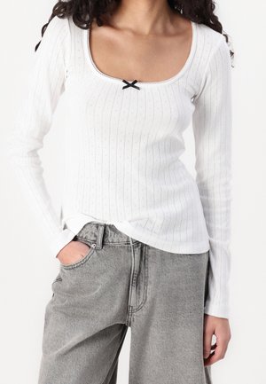 Witte geribbelde langemouwen top met een vierkante halslijn, een zwarte strikdetail in het midden en een licht gebogen zoom. Gecombineerd met grijze jeans.