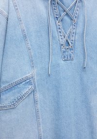 Top in denim blu chiaro caratterizzato da un scollatura con lacci, dettagli delle cuciture verticali e una tasca laterale sinistra. La texture è liscia con cuciture visibili.