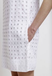 Robe blanche avec motifs carrés découpés, manches courtes et poches latérales. Confectionnée en tissu texturé au toucher léger.