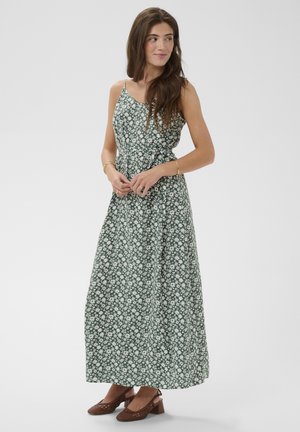 Kvinde iført en grøn ærmeløs maxikjole med hvidt blomsterprint og brune sko med lav hæl, stående med hænderne foldet og kigger til siden.