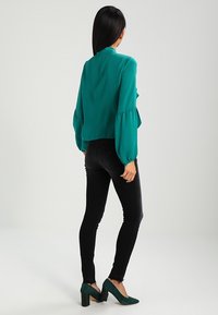 Blusa turquesa con mangas abullonadas, combinada con jeans ajustados negros y zapatos de tacón en bloque de ante verde, vista desde atrás.
