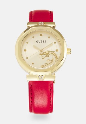 Guess-armur i guldfarve med rød læderrem, rund urskive med dragedesign, krystal-accenter og minimalistiske gyldne timeindikatorer.