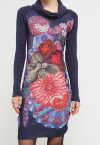 Långärmad klänning med cowl-urringning, blommigt mönster i röda, rosa och blå nyanser, med en texturerad mörkblå tyg på ärmarna och bakdelen.