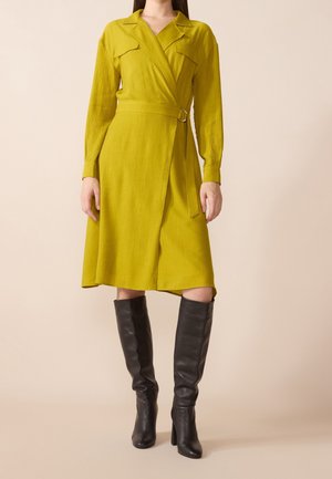 Kobieta ubrana w musztardowożółtą sukienkę kopertową z długimi rękawami i paskiem, połączoną z czarnymi skórzanymi botkami za kolano.