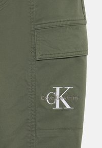 Pantalons cargo vert olive en mélange de coton avec une poche patch et un logo "CK" brodé en blanc. Texture lisse et coutures solides.