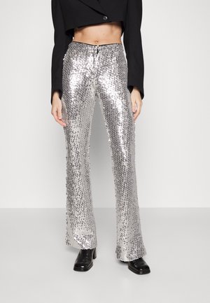 Personne portant un pantalon argenté à sequins taille haute, un top noir court à manches longues et des bottines noires sur fond blanc uni.