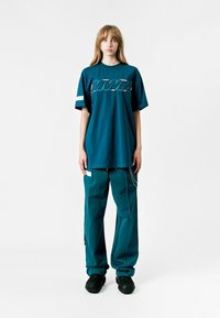 Camiseta oversized color teal con logo y detalles en blanco, combinada con pantalones teal a juego y calzado negro texturizado. Diseño casual.