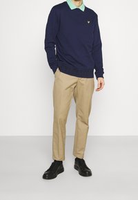 Suéter navy con camisa de cuello verde debajo, pantalones beiges y zapatillas de cuero negras, con un detalle de logo y texto amarillos en el dobladillo.