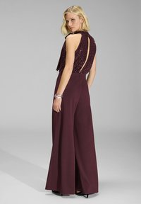 One-piece bordeaux con top in paillettes con scollatura sul retro, pantaloni larghi e finitura in tessuto liscia. Gli accessori includono un bracciale a più fili.