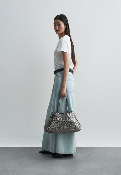 Υφασμένη μεταλλική γκρι τσάντα χειρός με δύο χερούλια, συνδυασμένη με λευκό t-shirt και ανοιχτού μπλε τζιν μακριά φούστα, σε απλό φόντο.