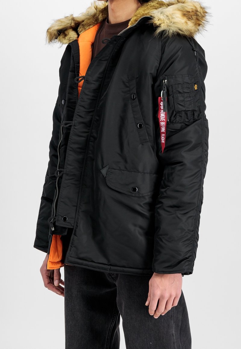 Parka negra aislante con capucha forrada de piel, forro naranja, cierre frontal con cremallera, múltiples bolsillos y una etiqueta decorativa en la manga.